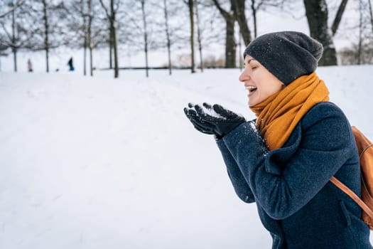 A young woman in warm winter clothes laughs looking at the snow in her palms