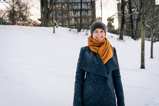 Smiling woman in a winter coat, scarf and hat against the backdrop of a winter snowy city.