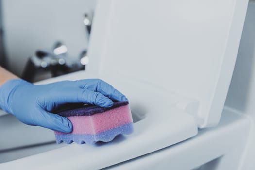 Woman hand closeup gloved in rubber protective gloves. Housewife cleaning toilet bowl, seat with detergent, pink cloth in bathroom or public restroom. Disinfection, hygiene, cleaning service concept