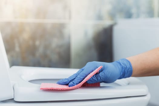 Cropped image view with close up on woman hands gloved in rubber protective gloves. Housewife cleaning toilet bowl, seat with pink cloth, wipe in bathroom or modern public restroom with blue grey tile