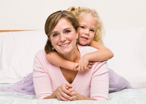 A mother is lying with her daughter on her bed.  The girl has her arms aroung her mother and they are smiling at the camera.  Horizontally framed shot.
