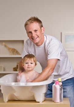 A young father is giving his baby daughter a bath in a childs bathtub.  He is smiling at the camera.  Vertically framed shot.