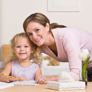 A mother is sitting with her young daughter and watching her draw.  They are smiling at the camera.  Square framed shot.