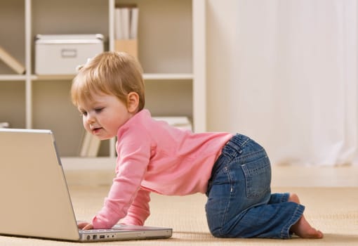 A baby girl is crawling next to a laptop.  She is looking at the screen.  Horizontally framed shot.