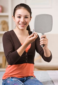 A young girl is looking into a handheld mirror and putting makeup on.  She is smiling at the camera.  Vertically framed shot.