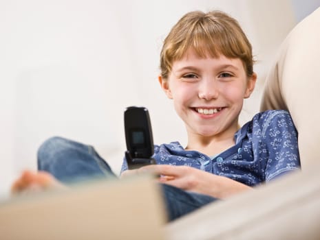 A young girl is holding a cellphone and smiling at the camera.  Horizontally framed shot.
