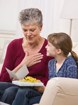 A young girl is handing a present to her grandmother, who is reading the card.  They are smiling and looking away from the camera.  Vertically framed shot.
