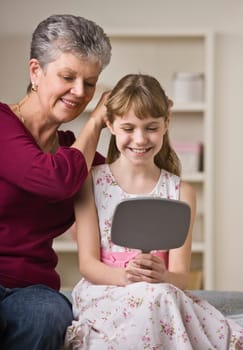A grandmother is sitting with her granddaughter who is looking at herself in the mirror.  They are smiling and looking away from the camera.  Vertically framed shot.