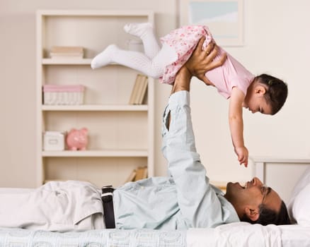 A father is holding his young daughter in the air above him.  They are smiling at each other.  Square framed shot.