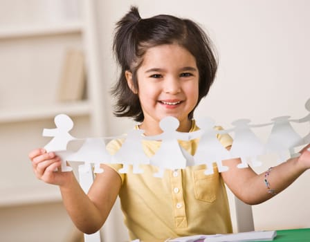 A young girl is seated at a desk and is holding up paper dolls.  She is smiling at the camera.  Horizontally framed shot.