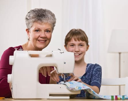 A young girl is sewing with her grandmother in the living room.  They are smiling at the camera.  Square framed shot.