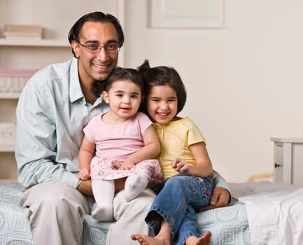 A father is sitting on a bed with his two young daughters.  They are smiling at the camera.  Horizontally framed shot.