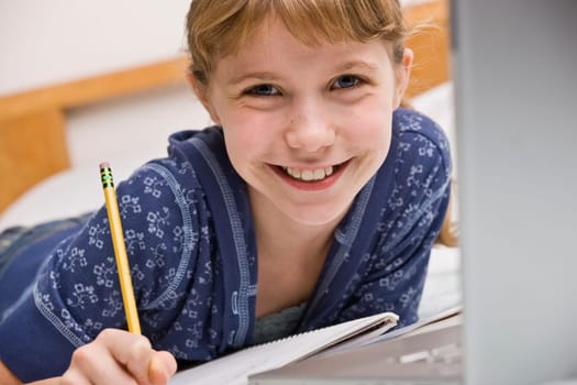 A young girl is seated at a computer desk and is working on her homework.  She is smilng at the camera.  Horizontally framed shot.