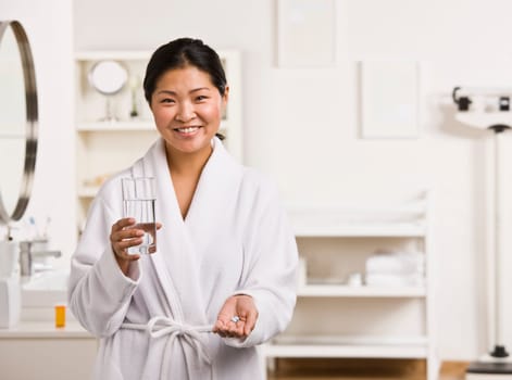 A woman is standing in a bathroom, and is about ot take some medication.  She is smiling at the camera.  Horizontally framed shot.