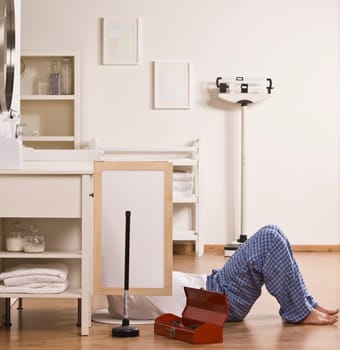 A man is on the floor in his bathroom, and he is fixing the kitchen sink.  Square framed shot.