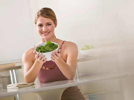 A young woman is seated in the kitchen and is holding out a bowl of salad.  She is smiling at the camera.  Horizontally framed shot.