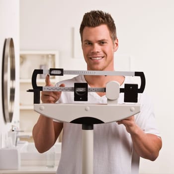 A young man is weighing himself on a bathroom scale.  He is smiling at the camera.  Square framed shot.