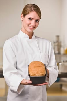 A young woman is standing in a kitchen and holding out a loaf of bread.  She is smiling at the camera.  Vertically framed shot.