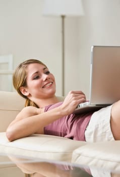 A young, smiling woman is laying on a couch and looking at a laptop.  Vertically framed shot.