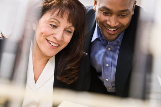 A businessman and woman are working together in an office.  They are smiling and looking away from the camera.  Horizontally framed shot.