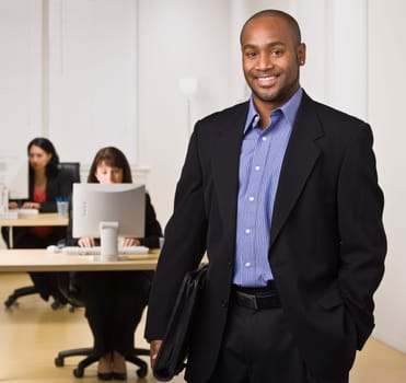 A young businessman is standing in an office with some other business people.  He is smiling at the camera.  Horizontally framed shot.