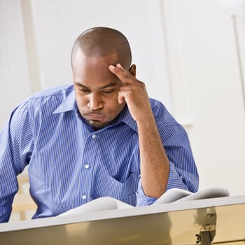 An attractive African-American male studying at a desk. He is resting his head against his fingers, and is puffing his cheeks out with air.  Square composition.