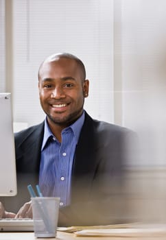 A young businessman is seated at a desk in front of a computer, and is smiling at the camera. Vertically framed shot.