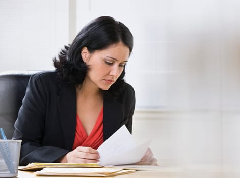 A young, attractive woman is looking over some paperwork on a desk.  She is looking away from the camera.  Horizontally framed shot.