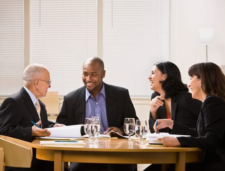 A group of business people are in a meeting in an office.  They are talking and laughing and looking away from the camera.  Horizontally framed shot.