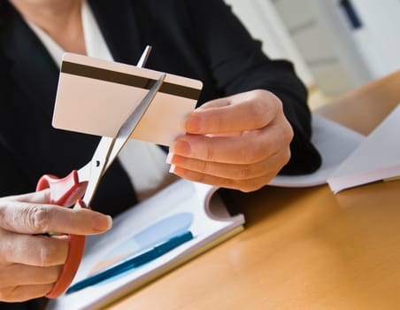 A businesswoman is seated at a desk and is cutting up a credit card with a pair of scissors.  Horiozntally framed shot.