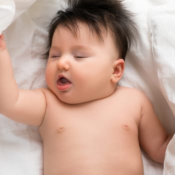An adorable baby lies on soft white bedding, peacefully asleep with eyes closed and an expression of calm happiness.