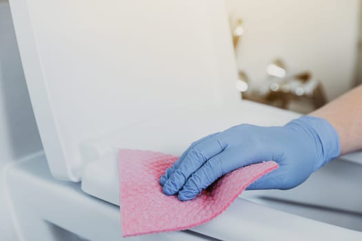 Close up on woman hand gloved in blue rubber protective gloves. Housewife cleaning toilet bowl, seat with detergent liquid, wet wipe in bathroom, restroom. Female washing, disinfecting toilet seat