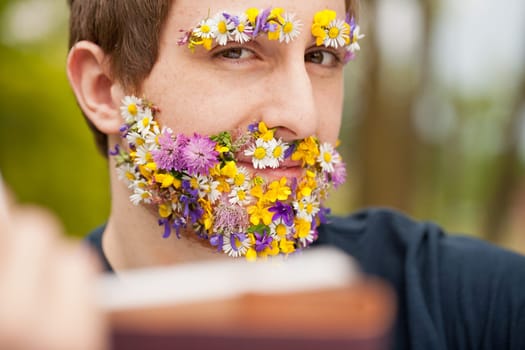 young man with his face flower-covered on his beard and eyebrows reading and staring at you very self-confident