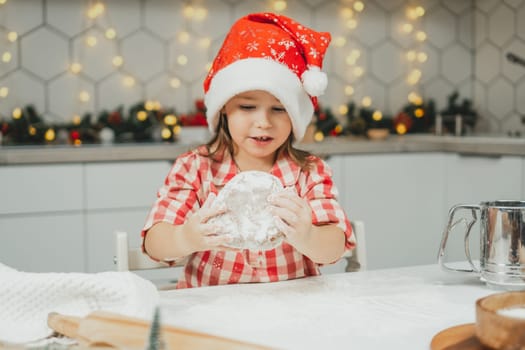 Little dark-haired girl 3 years old in red Christmas cap and checkered shirt prepares dough for gingerbread cookies in white Christmas decorated kitchen with garland lights. Merry Christmas, New Year