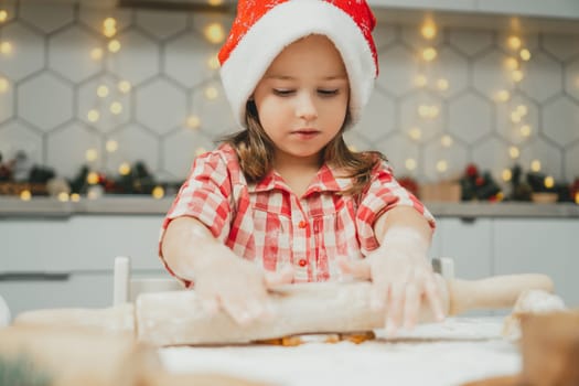 Little dark-haired girl 3 years old in red Christmas cap and checkered shirt roll out dough for gingerbread cookies in white Christmas decorated kitchen with garland lights. Merry Christmas, New Year