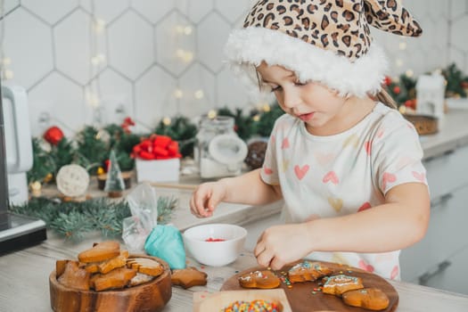 Little dark-haired girl 3 years old in Santa spotted cap decorate gingerbread cookies with icing in white Christmas decorated kitchen. Merry Christmas, Happy New Year