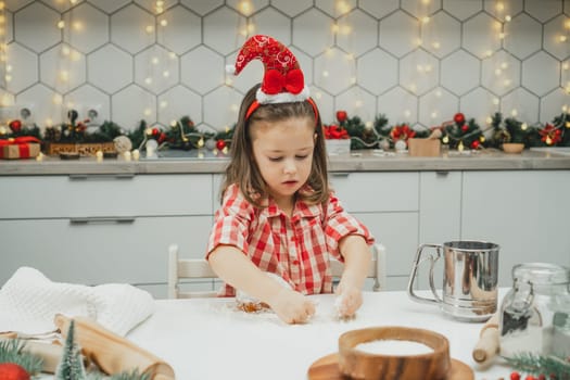 Little dark-haired girl 3 years old in red Christmas cap and checkered shirt prepares dough for gingerbread cookies in white Christmas decorated kitchen with garland lights. Merry Christmas, New Year