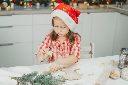 Little dark-haired girl 3 years old in red Christmas cap and checkered shirt cuts out gingerbread cookies from rolled dough in white Christmas decorated kitchen. Merry Christmas, Happy New Year