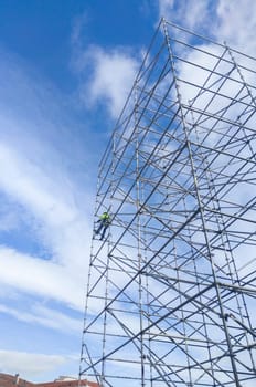 Unidentified worker at height dismantling a scaffolding tower. Blue cloudy sky on background