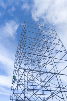 Unidentified worker at height dismantling a scaffolding tower. Blue cloudy sky on background
