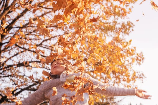 Partially blurred boy 9-10 years old stands in autumn park in rays of setting sun and throws up oak leaves, smiling and closing his eyes. Defocused child in gray sweater plays outdoors