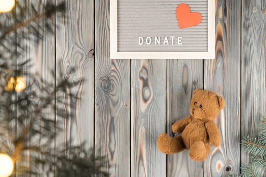 Brown teddy bear on gray wooden wall, top view. Social helping children concept. Decor from fir branches and garland lights. Giving money. New Year or Christmas charity, donation, kind heart.