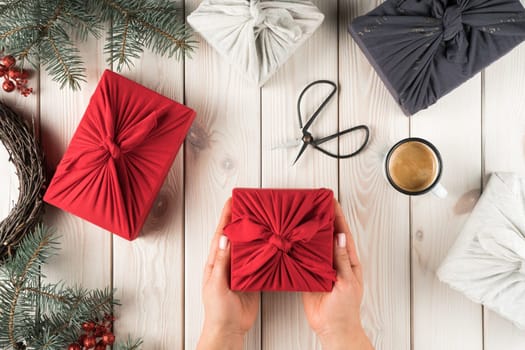 Female hands hold christmas present in furoshiki package, top view. Spruce branches, scissors, cup of coffee and gift boxes on planking. Christmas or New Year holiday concept, eco decor, zero waste.