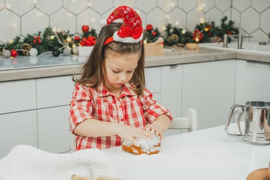 Little dark-haired girl 3 years old in red Christmas cap and checkered shirt prepares dough for gingerbread cookies in white Christmas decorated kitchen with garland lights. Merry Christmas, New Year
