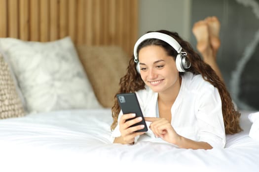 Woman using smart phone wearing headphones to listen to the music lying on the bed