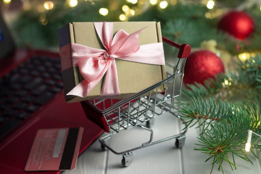 Shopping trolley with a gift near the keyboard of a red laptop, credit card, green branches of a Christmas tree with LED light bulbs, garland and shiny balls on a white wooden background. Merry Christmas and Happy New Year online shopping concept