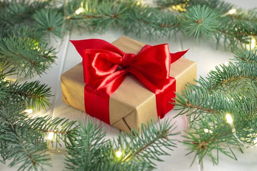 Gift wrapped in craft paper and red ribbon with green branches of a Christmas tree with LED light bulbs garland on a white wooden background. Merry Christmas and Happy New Year concept
