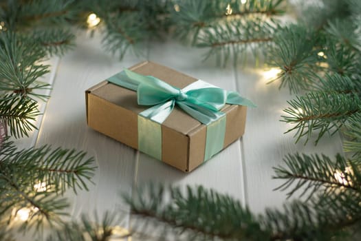 Gift wrapped in craft paper and mint ribbon with green branches of a Christmas tree with LED light bulbs garland on a white wooden background. Merry Christmas and Happy New Year concept