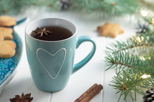 Mint cup of tea with heart, gingerbread, anise, cinnamon next to green branches of Christmas tree with LED light bulbs garland on a white wooden background. Merry Christmas and Happy New Year concept