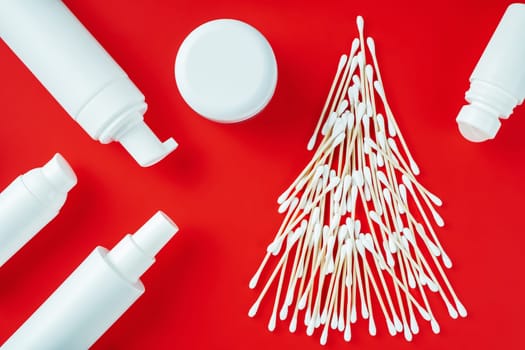 Wooden cotton swabs laid out in shape of Christmas tree with cosmetics jars and roll-on deodorant on red background. Flat lay